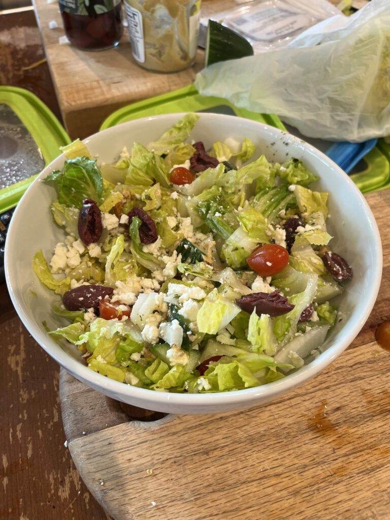 This is an image of a salad with romaine lettuce, tomatoes, kalamata olives, and feta. It is in a bowl on a wooden countertop.
