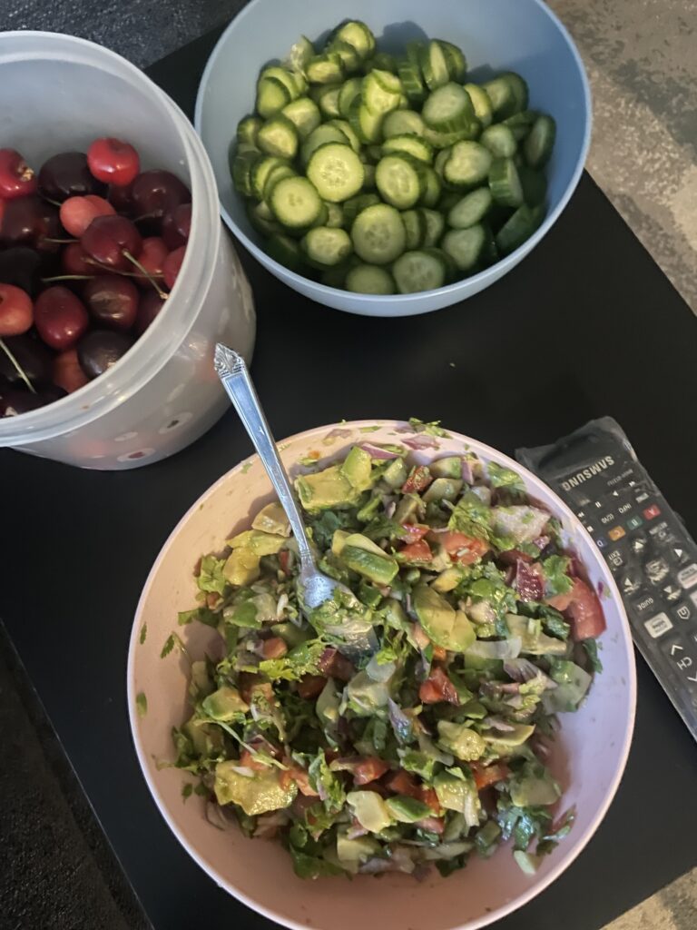 This is a photo of a mixed salad with tomato, avocado, cucumber, red onion, greens, and dressing. It is in a pink bowl with a spoon. Next to the salad, positioned above and to the left of it, are a bowl of sliced cucumbers, and a bowl of whole cherries.