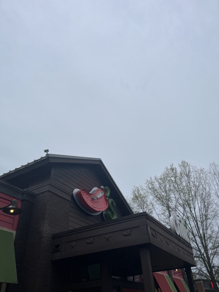 The upper exterior of the entrance of a Chili’s with a brown exterior accessed by green and red window awnings. There is a large Chili Pepper with a green apostrophe and letter S on the facade and the word “chilli’s” in white with the apostrophe in green on the entrance roof.