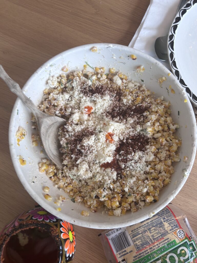 This is a picture of elote salad, a corn salad with a spicy and sour mayonnaise dressing and cojita cheese. The salad is in a white bowl with a large spoon.