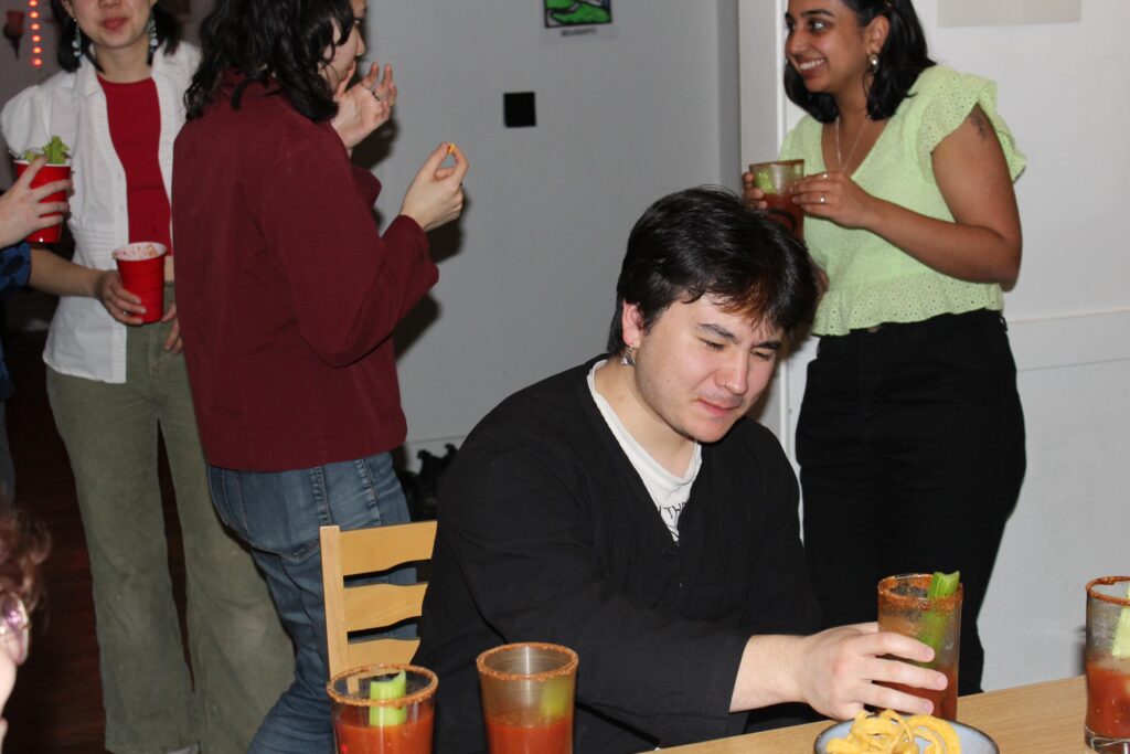 In a dining room, people are drinking bloody marys and enjoying each other’s company. Aidan Sweney takes a sip of his bloody mary and is repulsed.