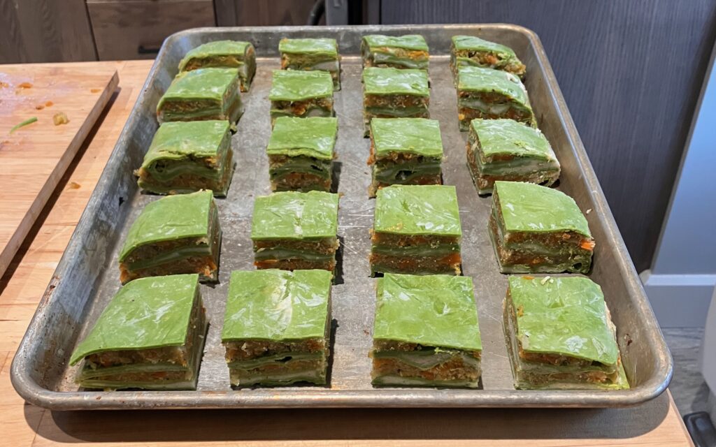 Twenty evenly sliced squares of raw green lasagna sit on a baking sheet.