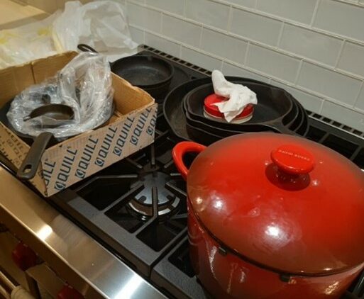 Ten skillets of assorted sizes and a Dutch oven sitting on the burners of a stove. The skillets are stacked and some are unwrapped hastily in packing material