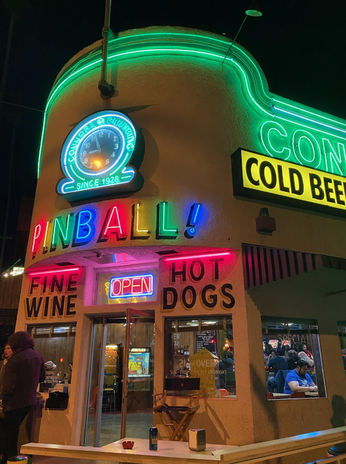 building with neon clock reading “Connor’s Plumbing Since 1926”, neon sign reading “Pinball!”, two painted signs reading “fine wine” and “hot dogs”