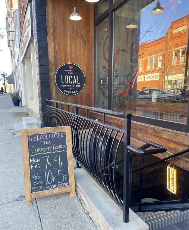 The entrance to a coffee shop from street level, with a black railing and a set of metal stairs descending until they disappear beneath the pavement. On the sidewalk there is a blackboard sign with the open hours. Facing the descending staircase is a circular black sign that says “The Local Coffee & Tea.”