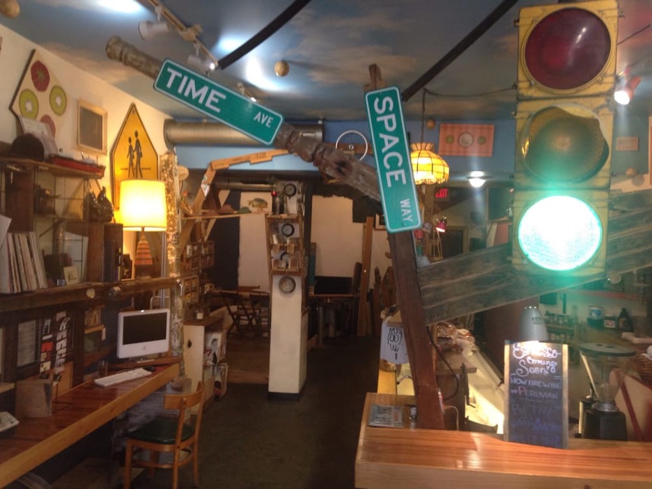 The dimly lit interior of Oh Yeah. The angular street post with “time ave” and “space way” street signs is the center of the frame. The ceiling is painted to look like a sky with fluffy white clouds. There are multiple clocks in view, a crosswalk sign, and both ceiling and table lamps. A street light lit up green shines on the right side of the image.