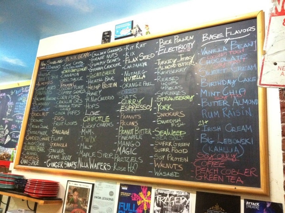 A blackboard with around eighty toppings written in alphabetical order with multicolored chalk, including balsamic vinegar, caramel, chocolate chips, cinnamon, corn pops, goat cheese, hemp protein, love, kit-kat, espresso, magic, rose h20, bee pollen, and electricity. Twelve base flavors are listed on the right hand side of the blackboard.